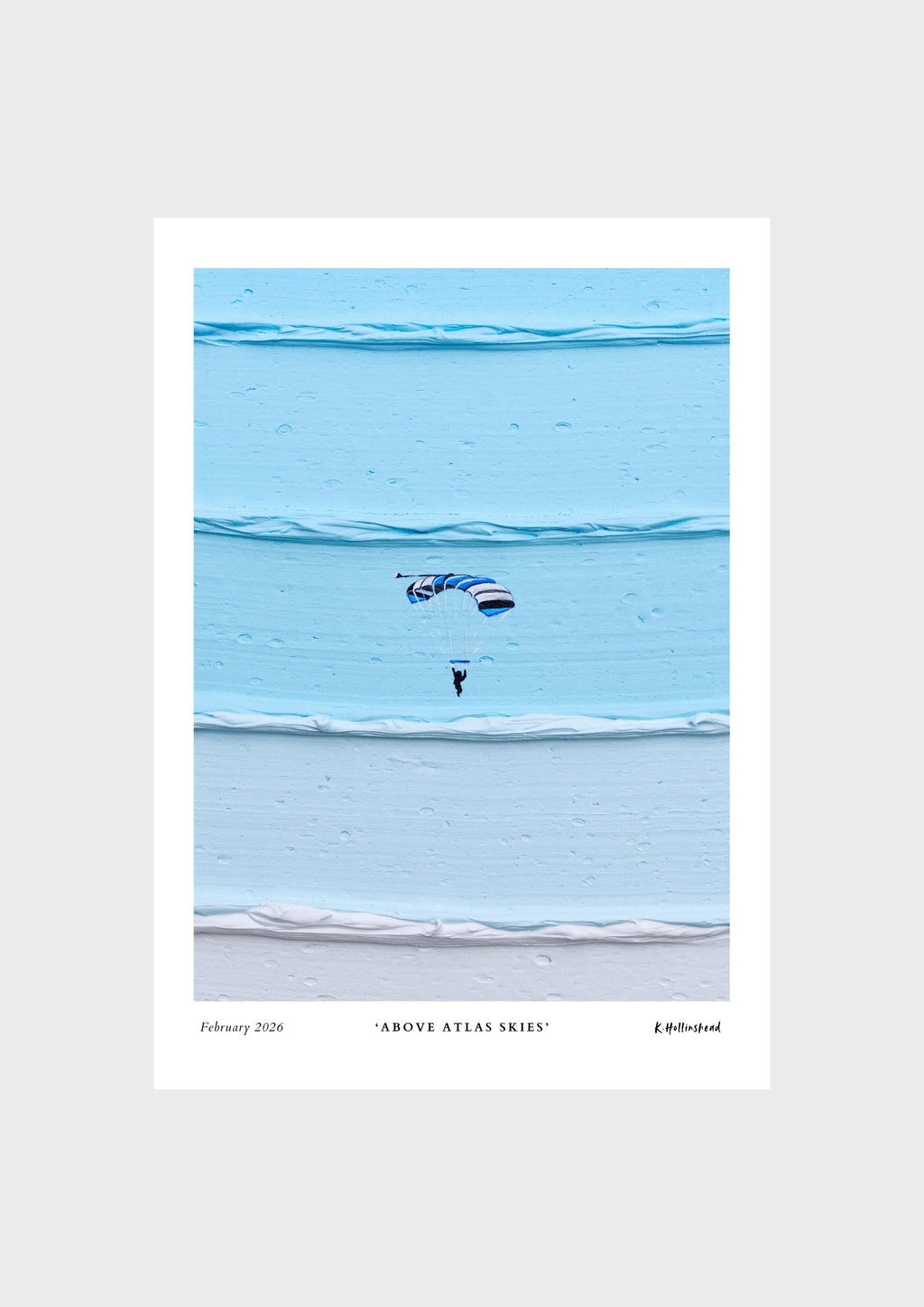 A minimalist art print titled 'Above Atlas Skies' featuring a small skydiver under a blue and white parachute against layered light blue water, presented on a light gray background.