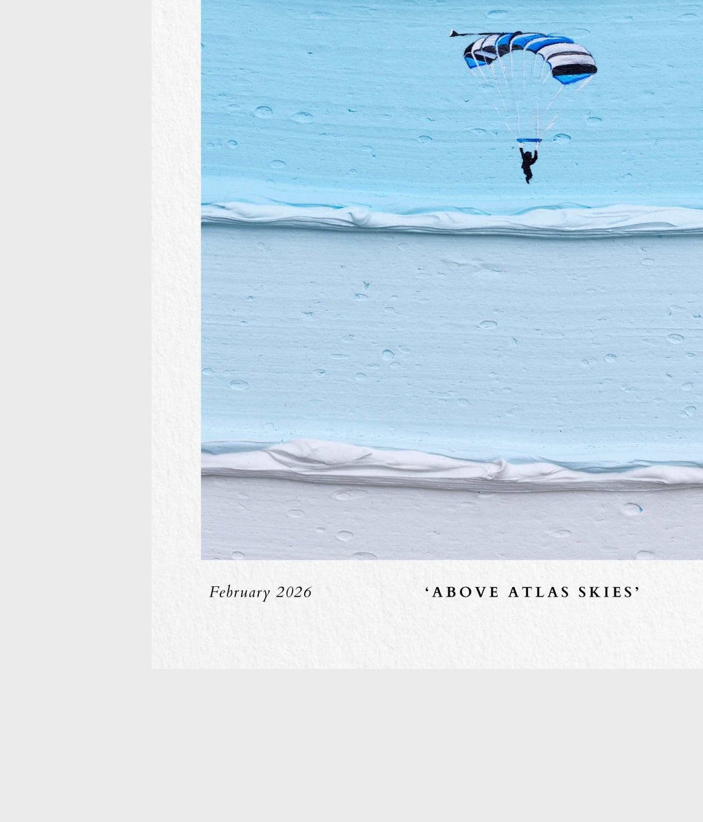 A square art print or poster featuring a minimalist photograph of a skydiver under a blue and white striped parachute over a light blue icy or snowy landscape. The print has a white border with the text 'February 2026' and '"ABOVE ATLAS SKIES"' printed at the bottom.
