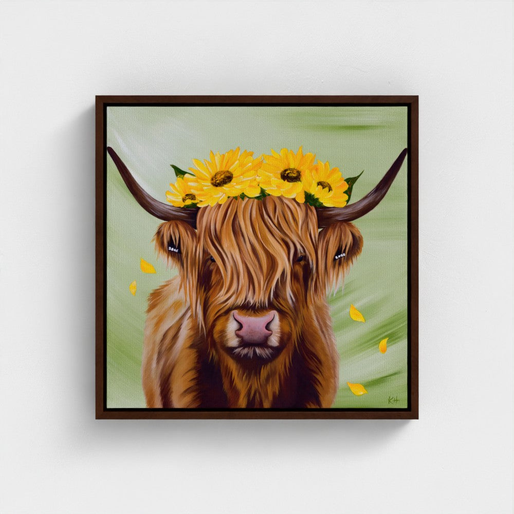 A framed square canvas print featuring a stylized painting of a Highland cow with long brown hair, a pink nose, and a crown of yellow sunflowers, displayed against a light gray wall.