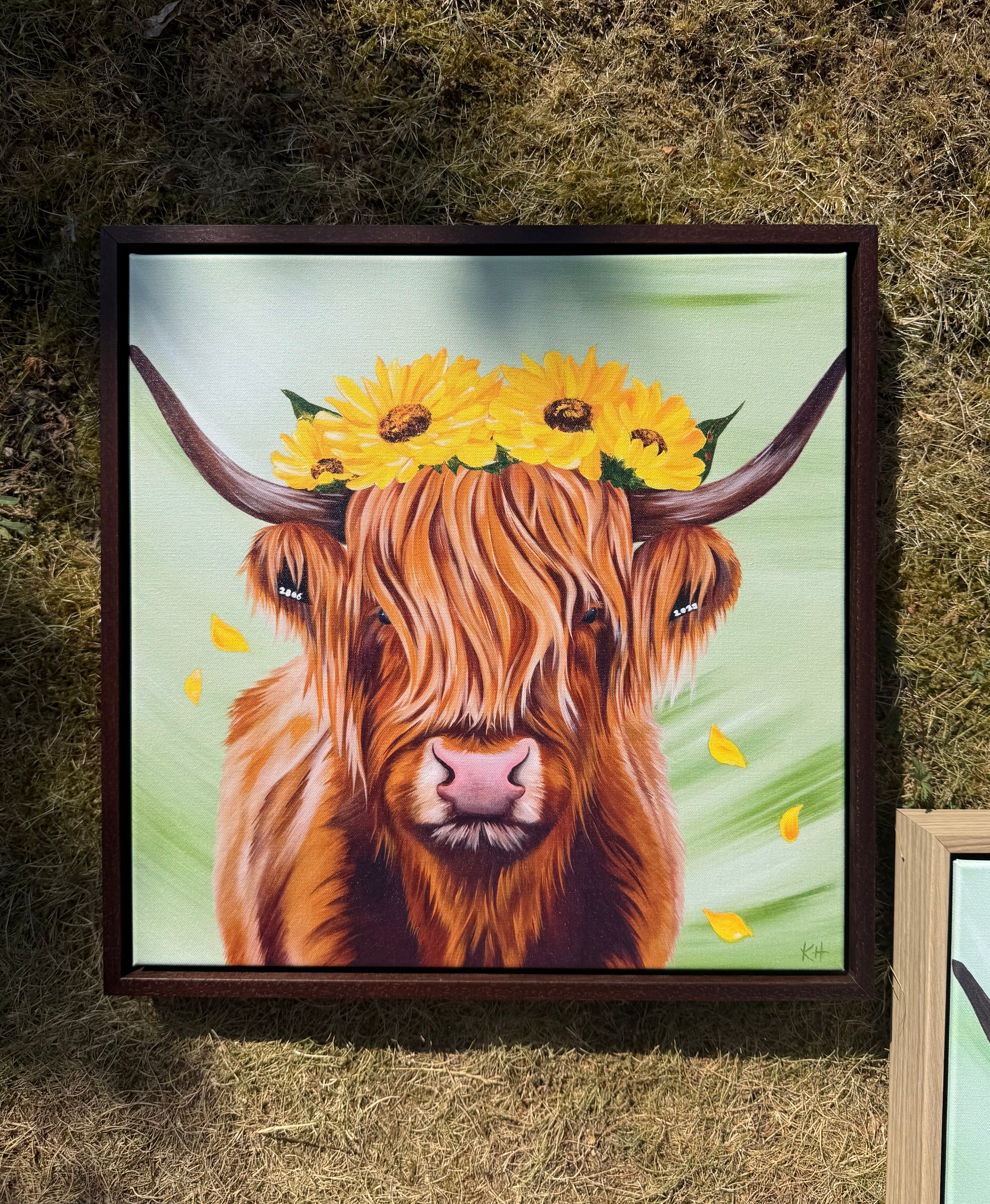 A framed painting of a Highland cow with yellow sunflowers on its horns, displayed on a bed of dry grass. The painting features a light green background with a few yellow leaves, and the cow is framed in a dark brown wooden frame.