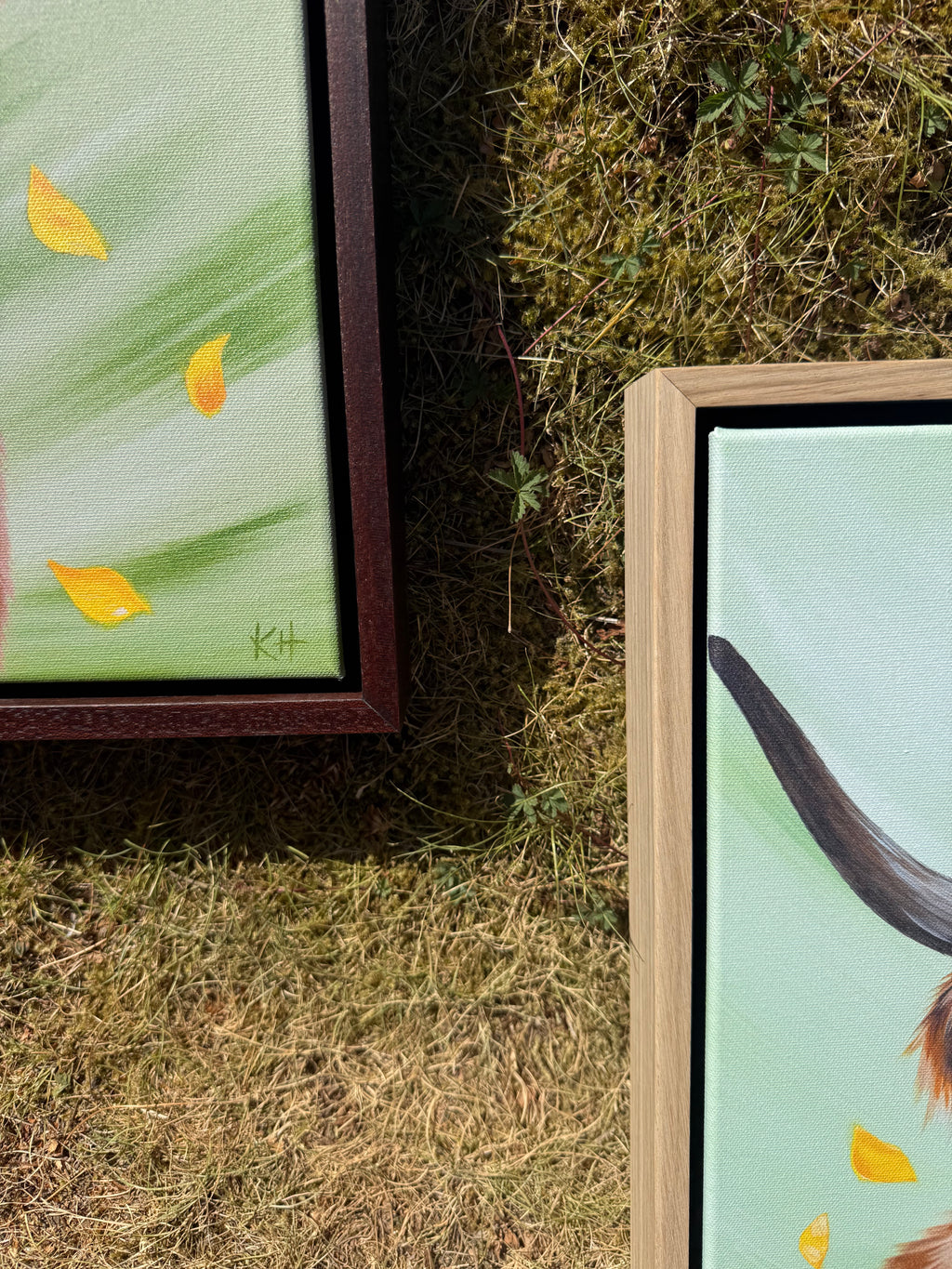 Two framed paintings displayed outdoors on grass. The left painting has a dark brown frame and shows yellow leaves on a light green background.