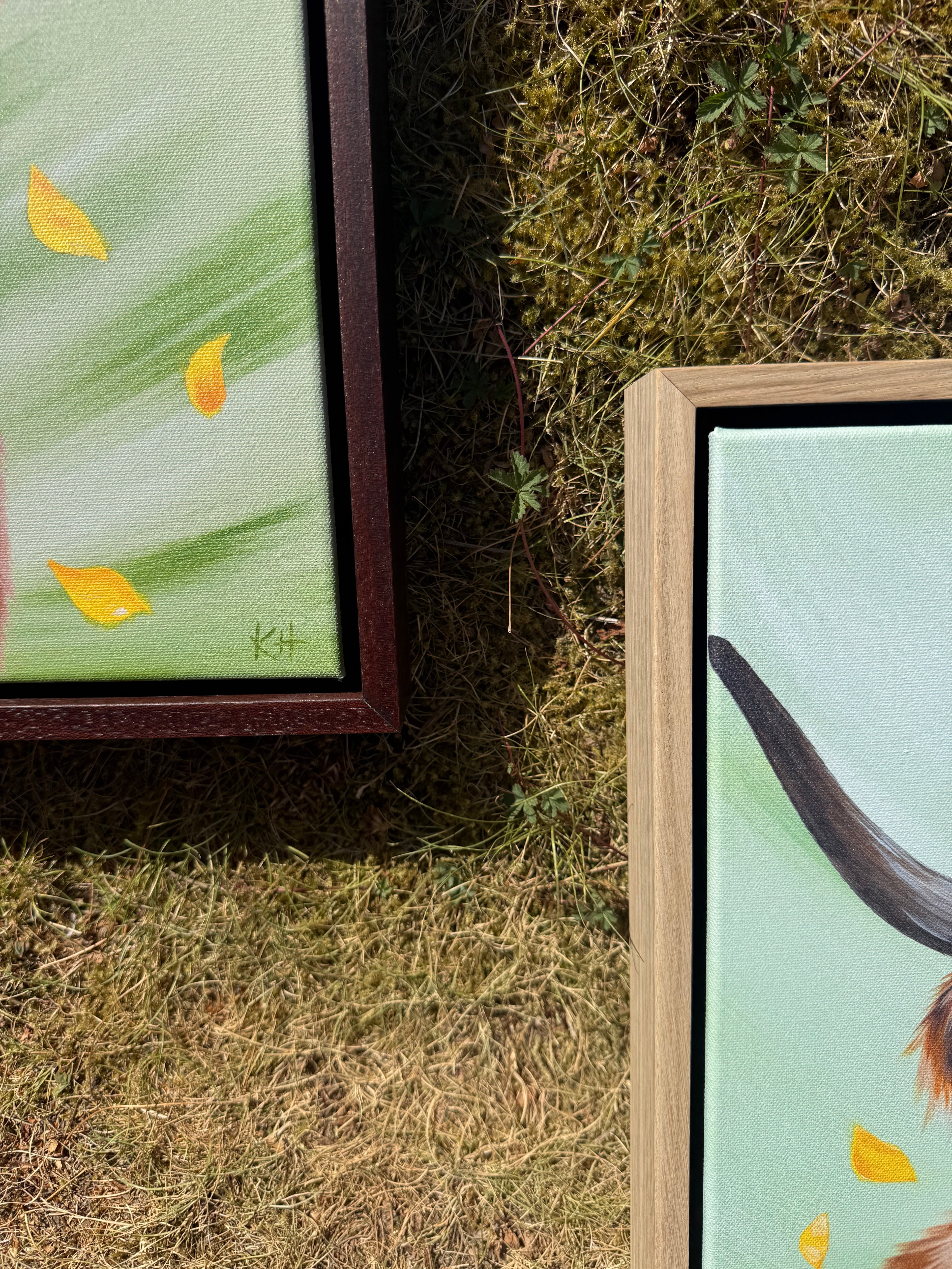 Two framed paintings displayed outdoors on grass. The left painting has a dark brown frame and shows yellow leaves on a light green background.