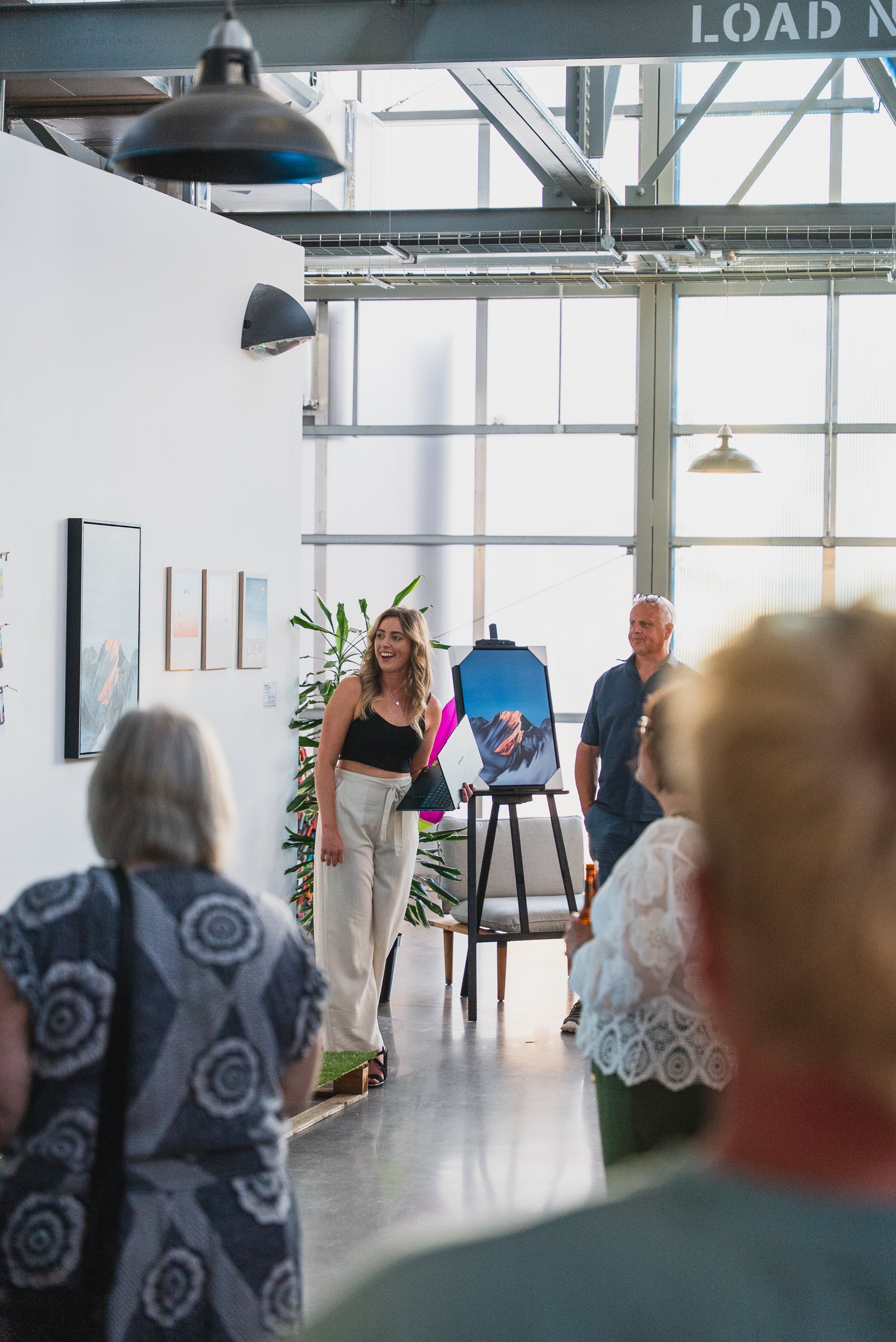 An indoor art gallery or studio event with several people visible, including a woman walking toward the camera near a framed artwork and an easel, with the backs of two audience members in the foreground.