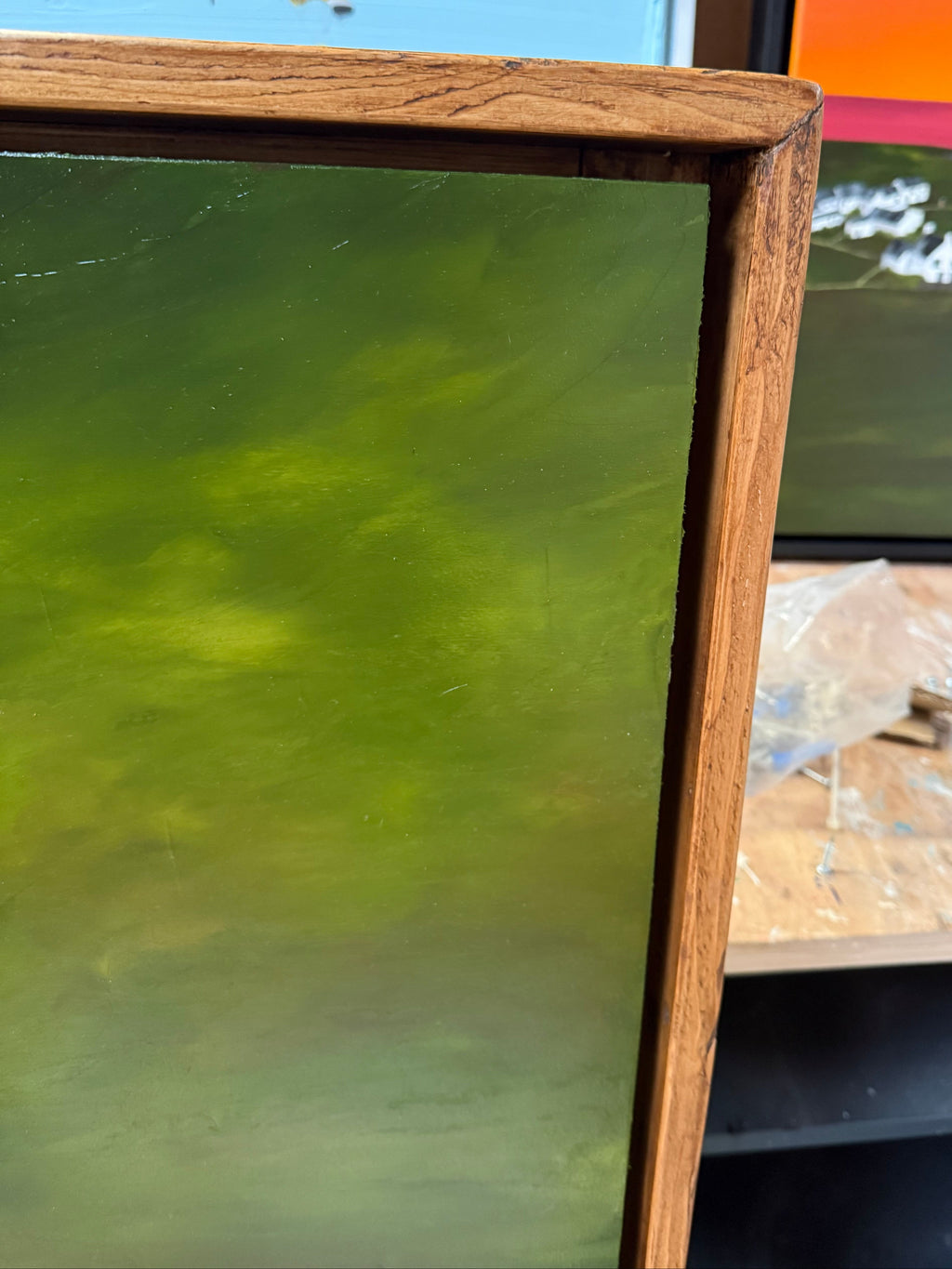 Close-up of a framed artwork showing a textured green painted surface with a medium-brown wooden frame, photographed indoors with other items partially visible in the background.
