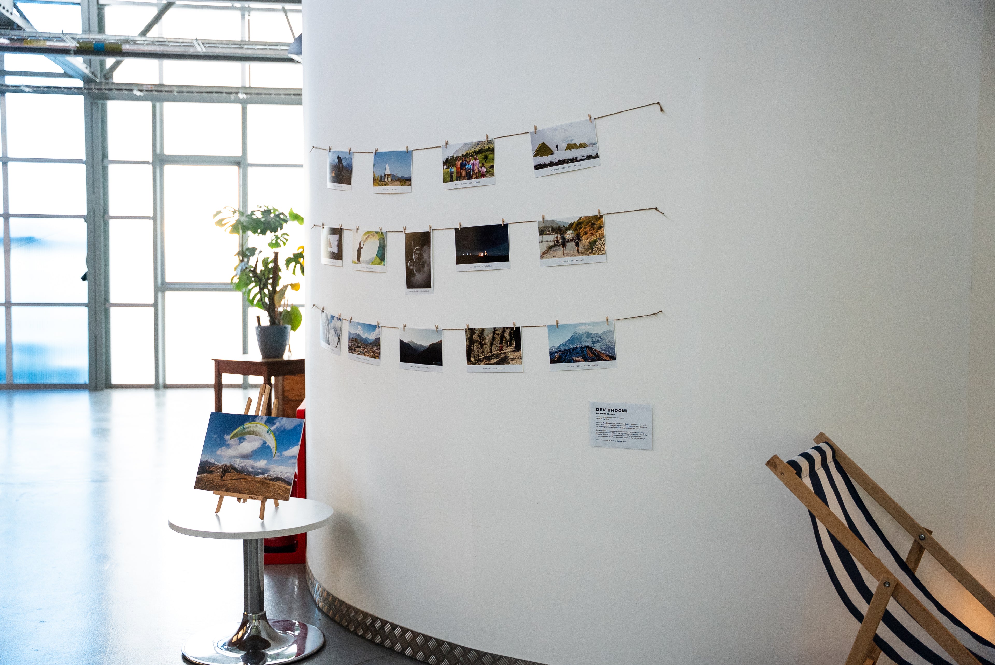 An indoor gallery or exhibition space with a curved white wall displaying multiple small landscape photographs strung across the wall. A small easel with a landscape print stands on a round table near a large window, and part of a wooden lounge chair is visible in the foreground.
