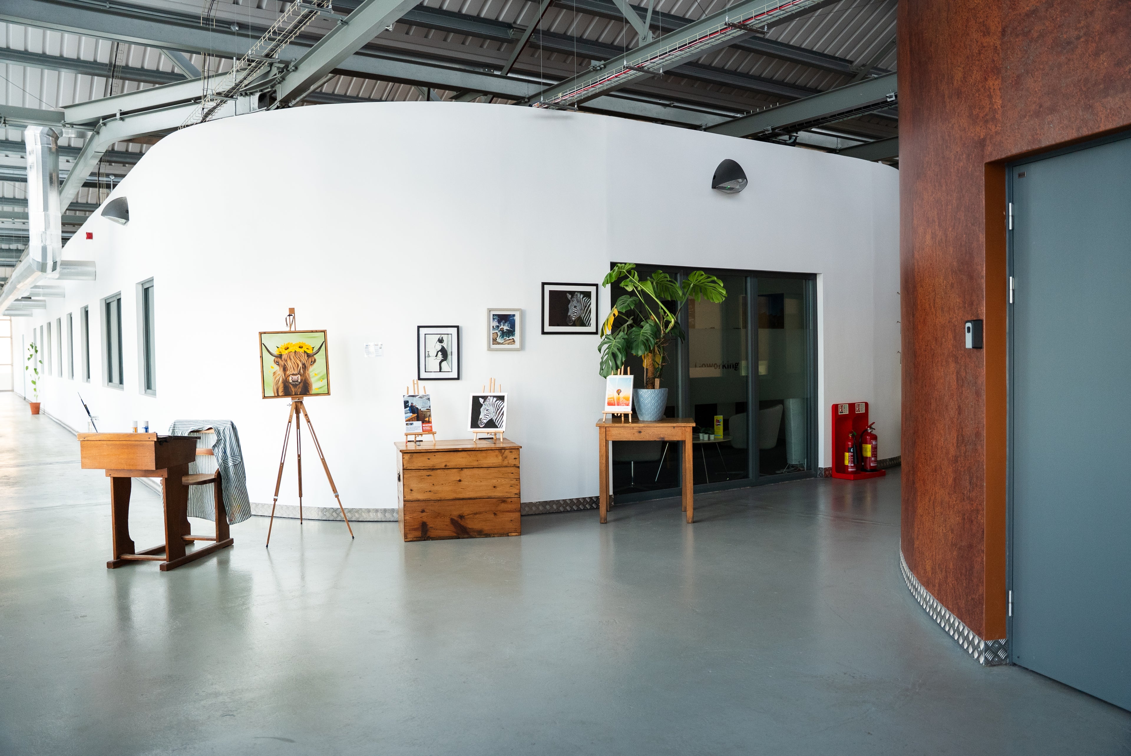 A wide interior hallway with a curved white wall, gray concrete floor, exposed metal ceiling, wooden furniture, framed wall art, a potted plant, and a closed gray door on the right.