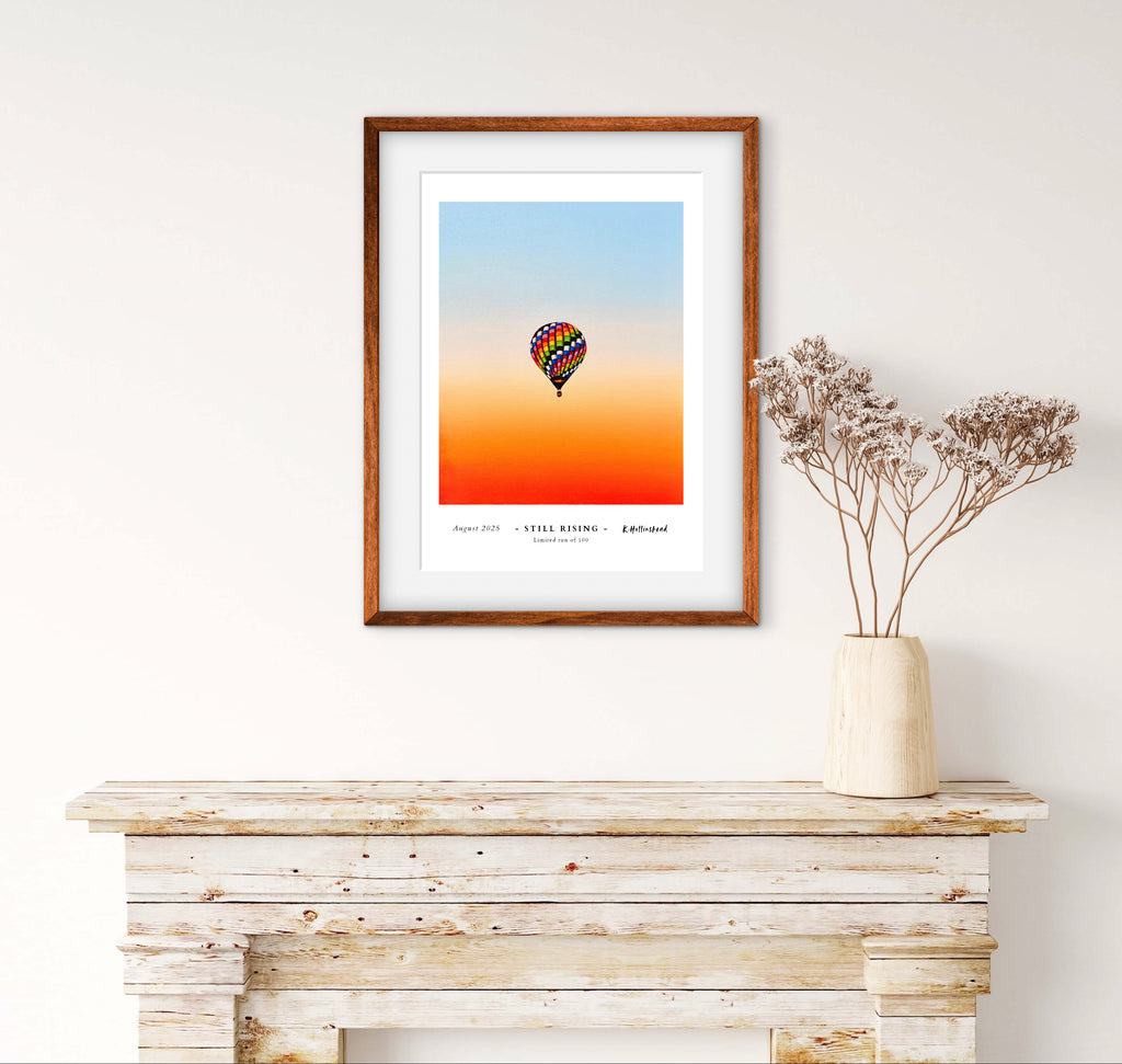 Framed artwork of a hot air balloon against an orange and blue gradient sky, displayed on a wooden surface.