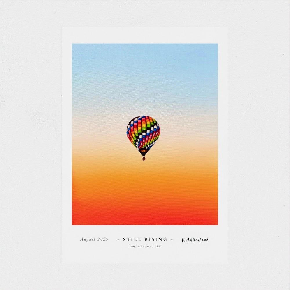 A square art print featuring a colorful hot air balloon against a gradient sky from light blue to orange and red. The print has a white border with printed text at the bottom reading 'August 2025 - STILL RISING - R.