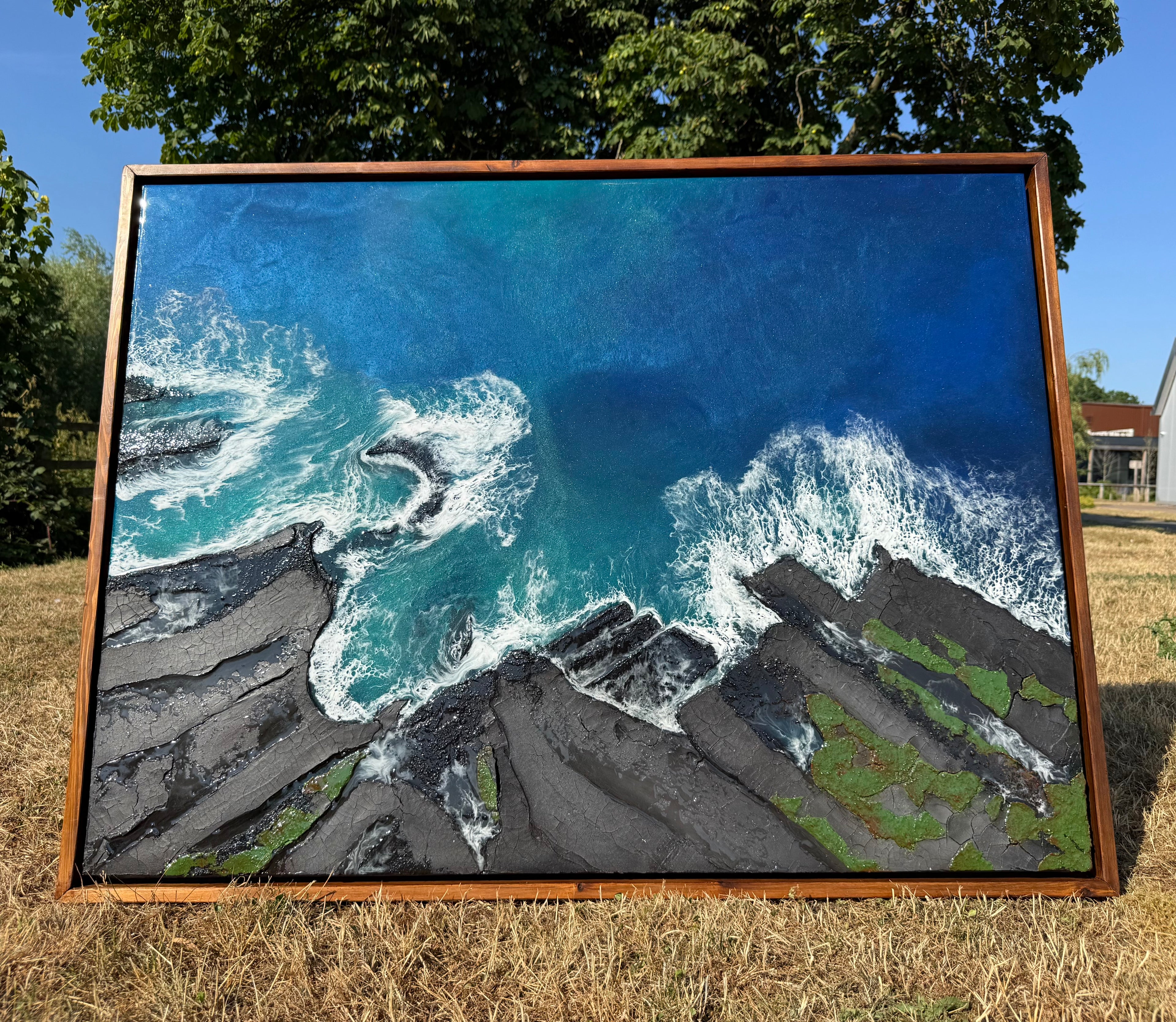 A framed landscape painting of resin ocean waves crashing on a rocky shore, displayed outdoors on dry grass with trees and a house in the background.