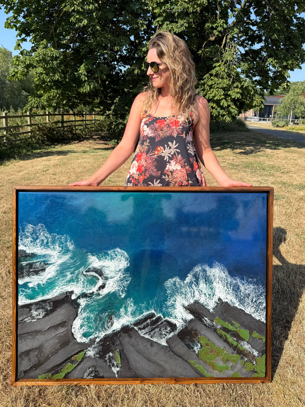 Katie Hollinshead wearing a floral dress and sunglasses stands outdoors in a grassy yard holding a large framed landscape painting of ocean waves crashing against dark rocky cliffs.