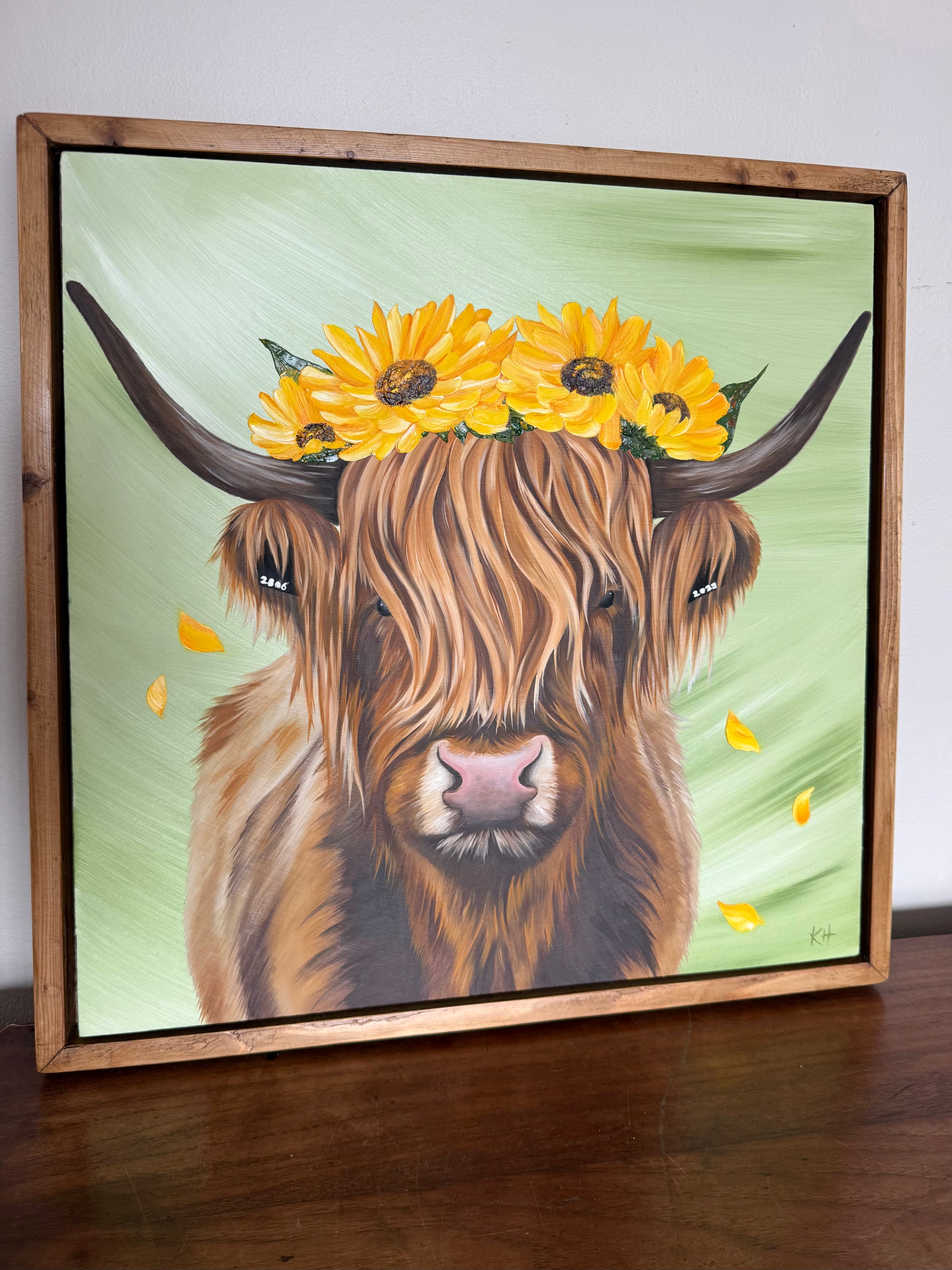A framed painting of a shaggy Highland cow with yellow sunflowers on its head, displayed on a wooden surface against a light wall.