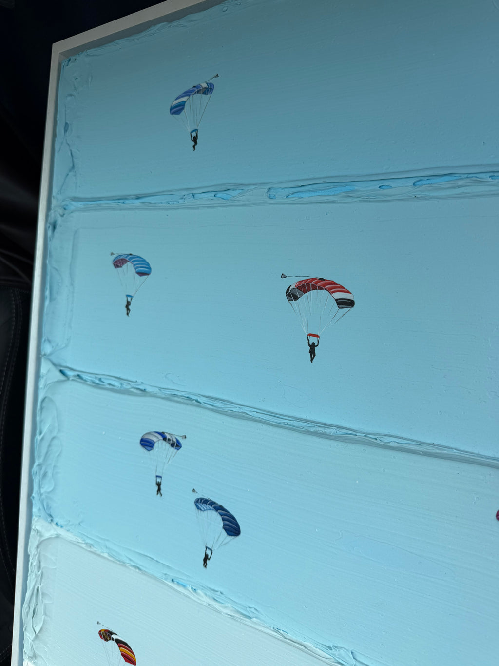A square glass art piece featuring a light blue sky background with multiple colorful parachutes in blue, red, and orange, framed by a white border.