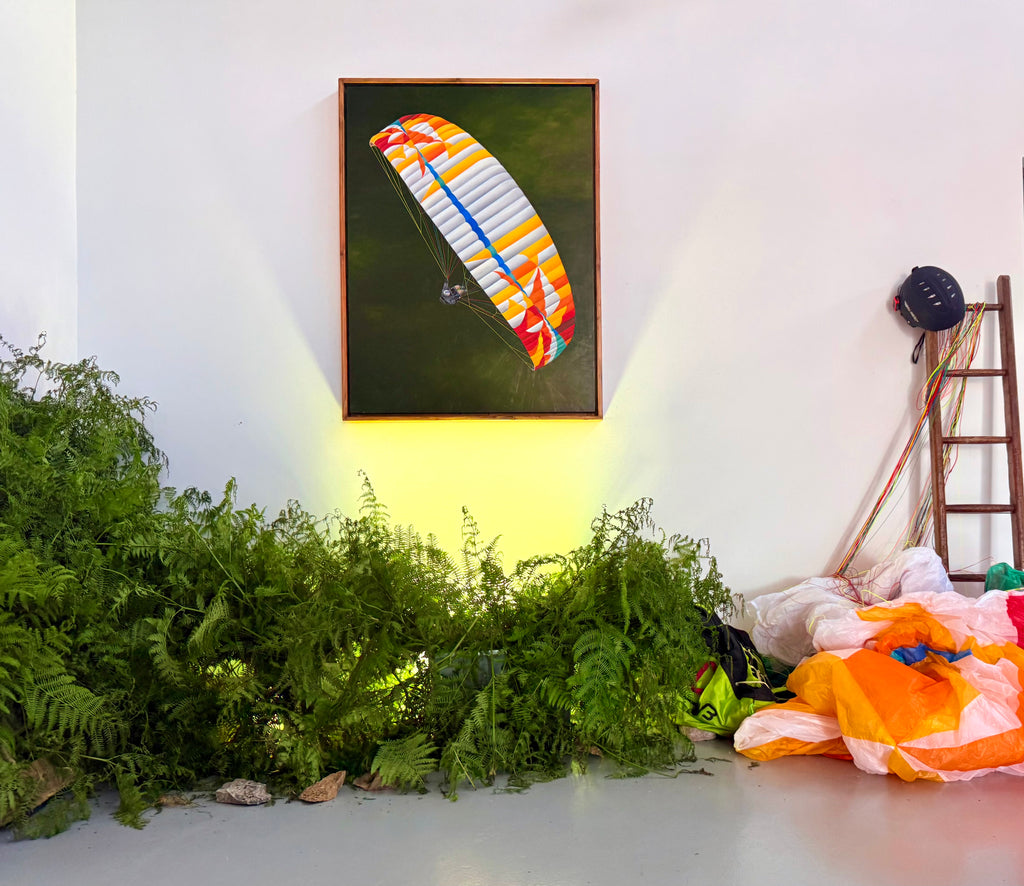 A framed original oil painting artwork depicting a colorful paraglider wing against a dark green background, displayed on a white wall in a gallery-like setting with a spotlight below it. The scene is decorated with green ferns along the floor, and a ladder, helmet, and orange/white paraglider fabric are visible on the right.