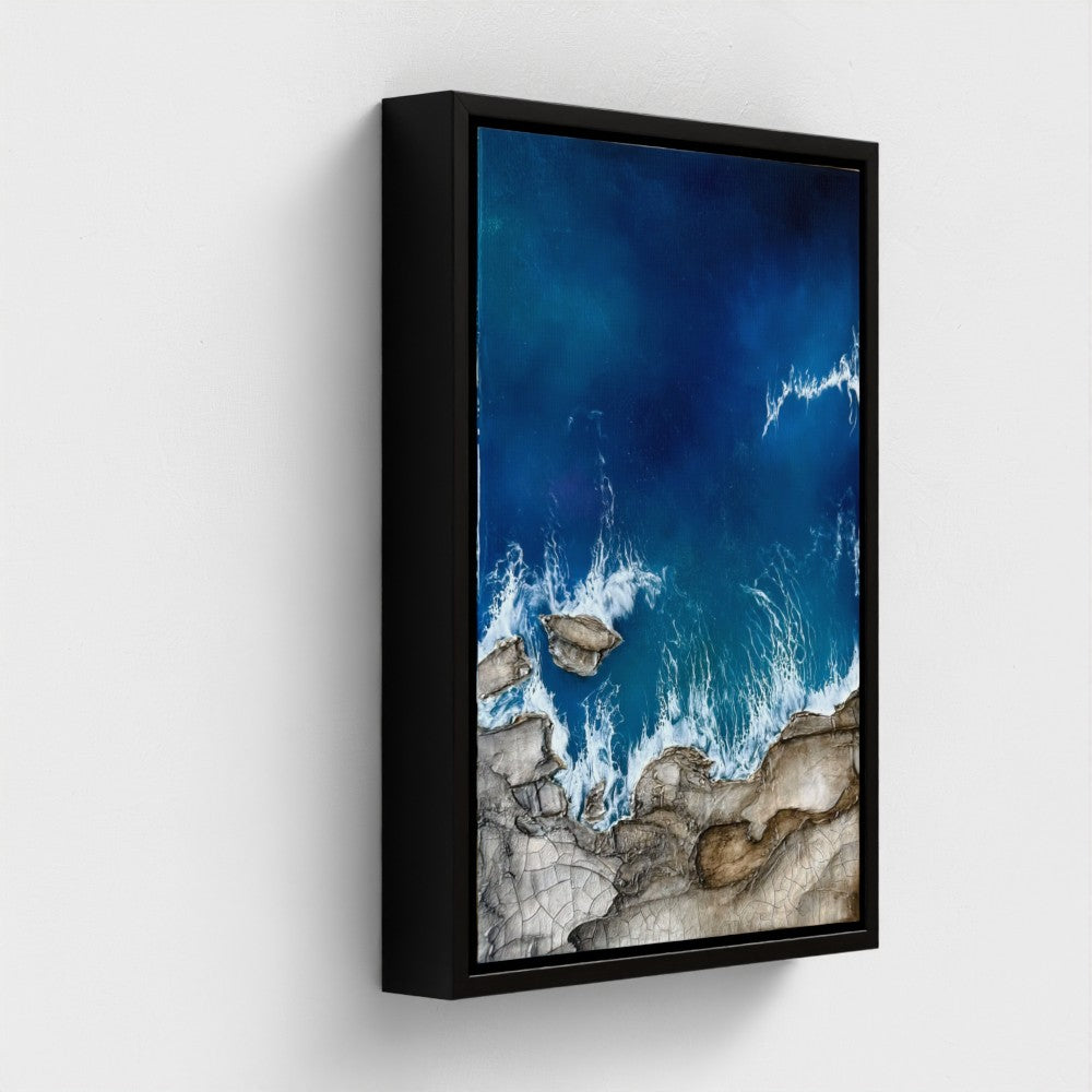 Framed artwork of a coastal scene with blue water and rocky shore on a white wall.