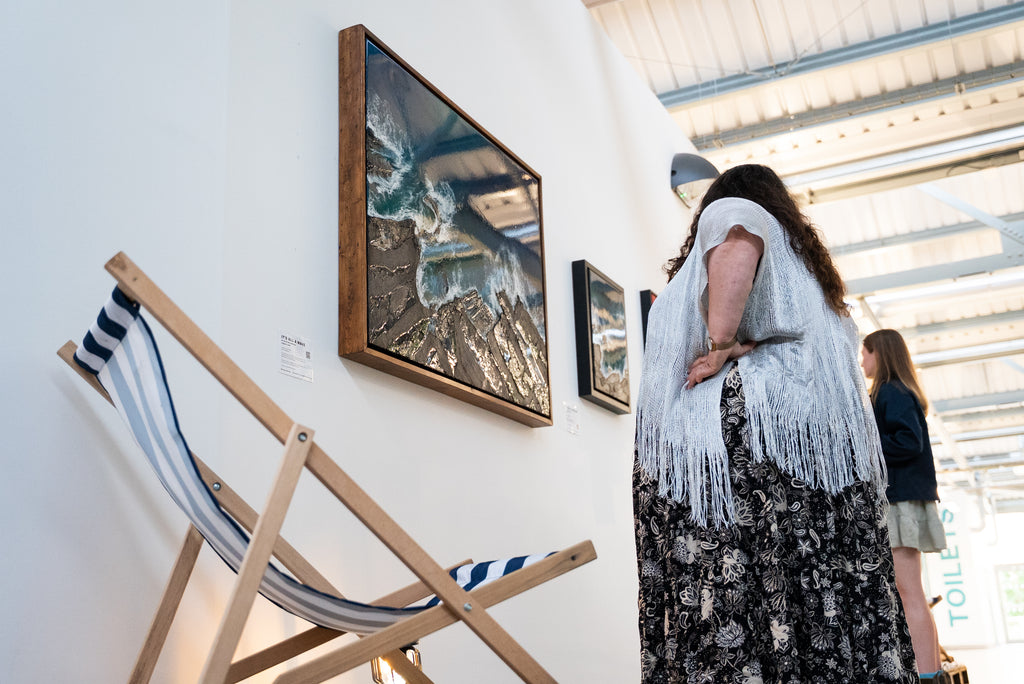 A woman in a white fringed cardigan and black floral dress stands in an art gallery, viewing a OCEAN RESIN WAVE ART PRINT on a white wall. A wooden deck chair with blue and white stripes is in the foreground, and another person is visible in the background.