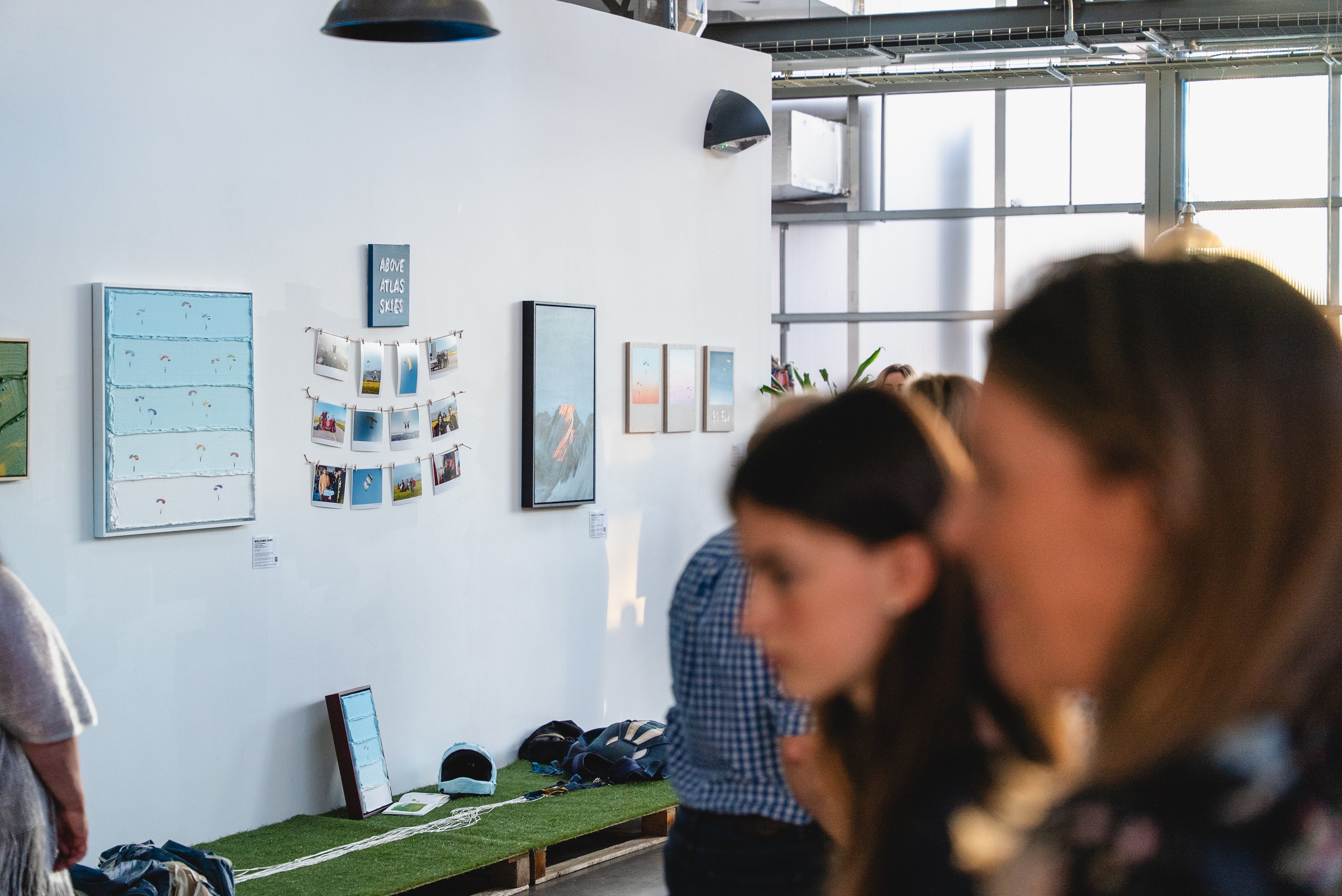 An indoor art gallery or studio space with several people in the foreground, a white wall with framed prints and a calendar, and a green bench along the floor.