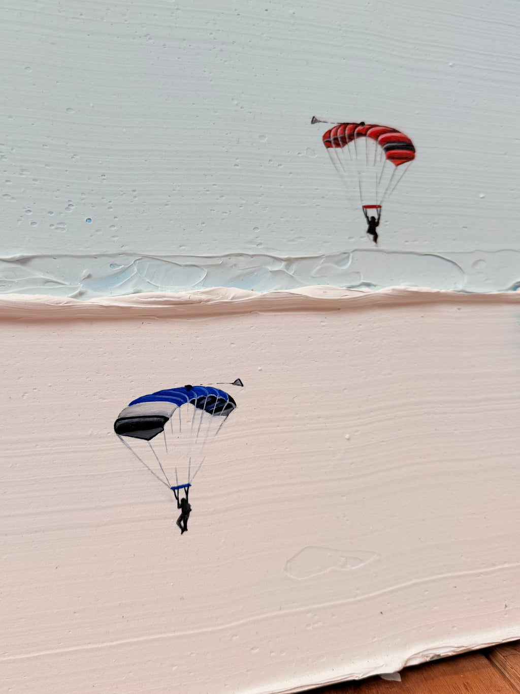 A hand-painted canvas print featuring two tiny parachutists floating in the sky, with one red and black striped parachute and one blue, black, and white parachute, set against a split white and beige background.