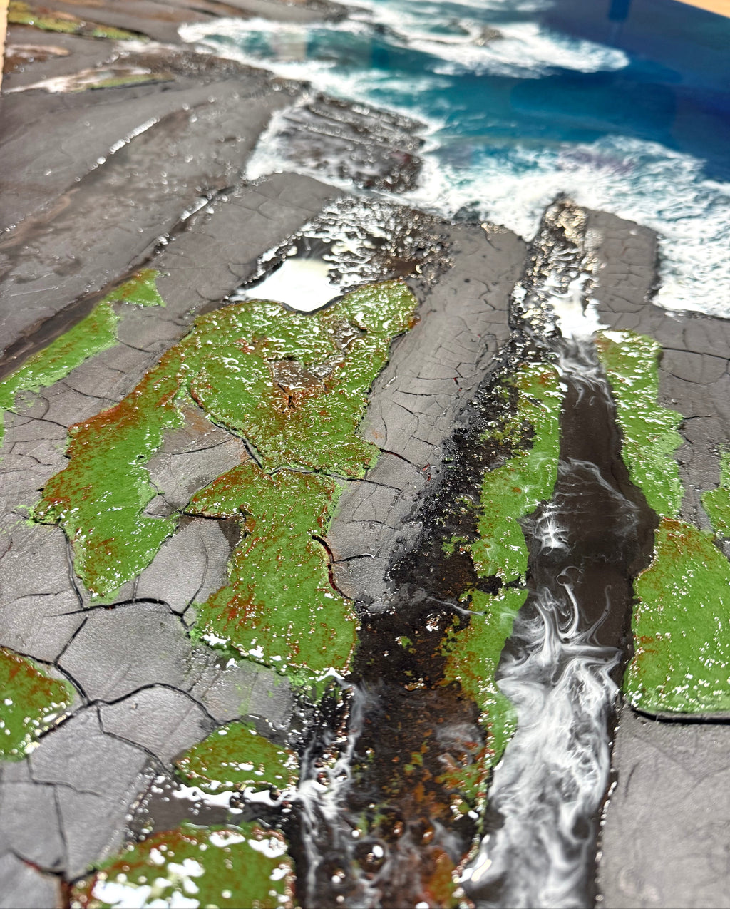A close-up of a textured coastal landscape artwork or print showing dark cracked ground, bright green mossy areas, white surf, and blue ocean water.
