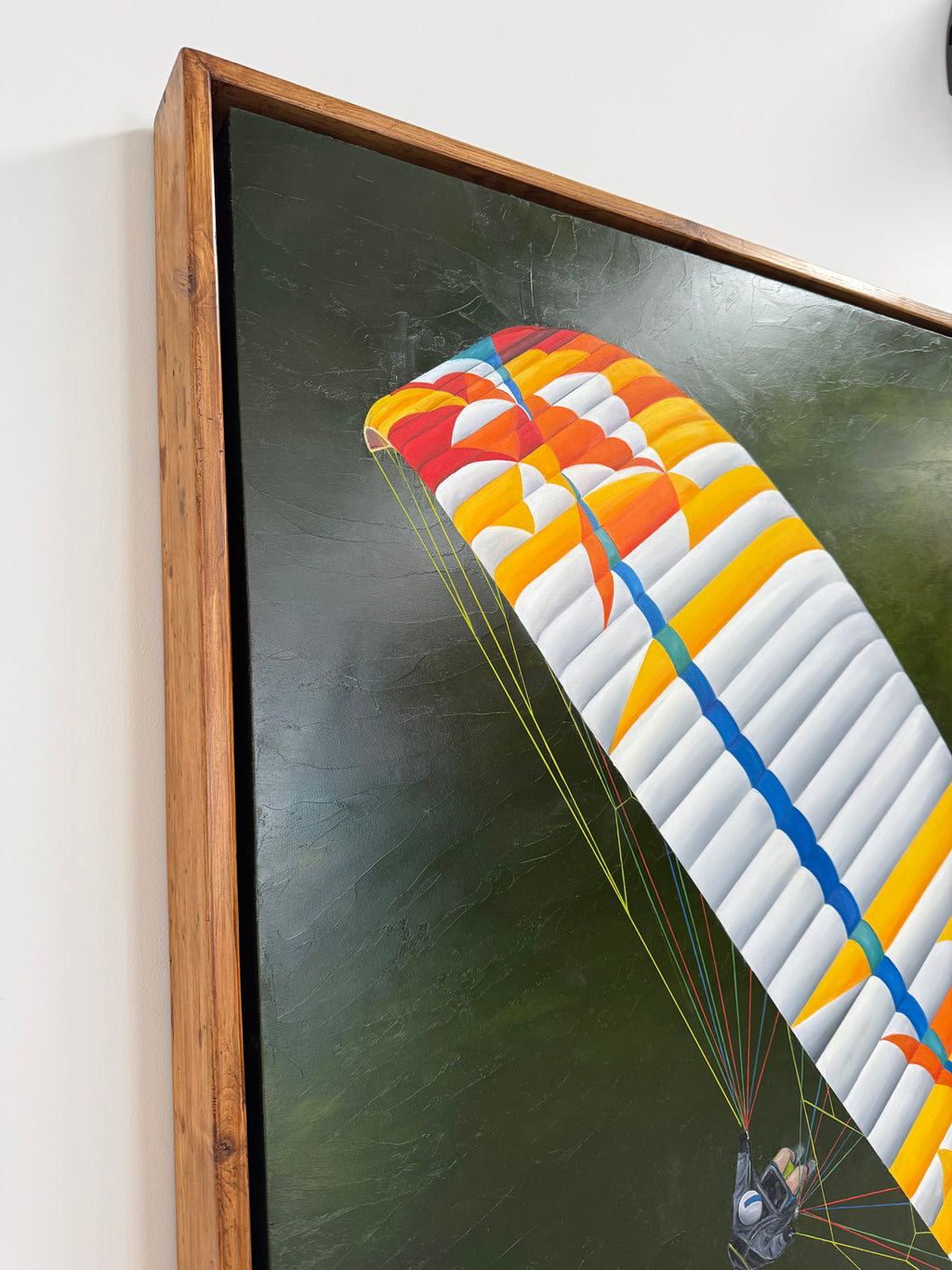 A framed artwork by katie hollinshead depicting a colorful paraglider canopy against a dark green background, shown hanging on a white wall. Handmade reclaimed wooden frame