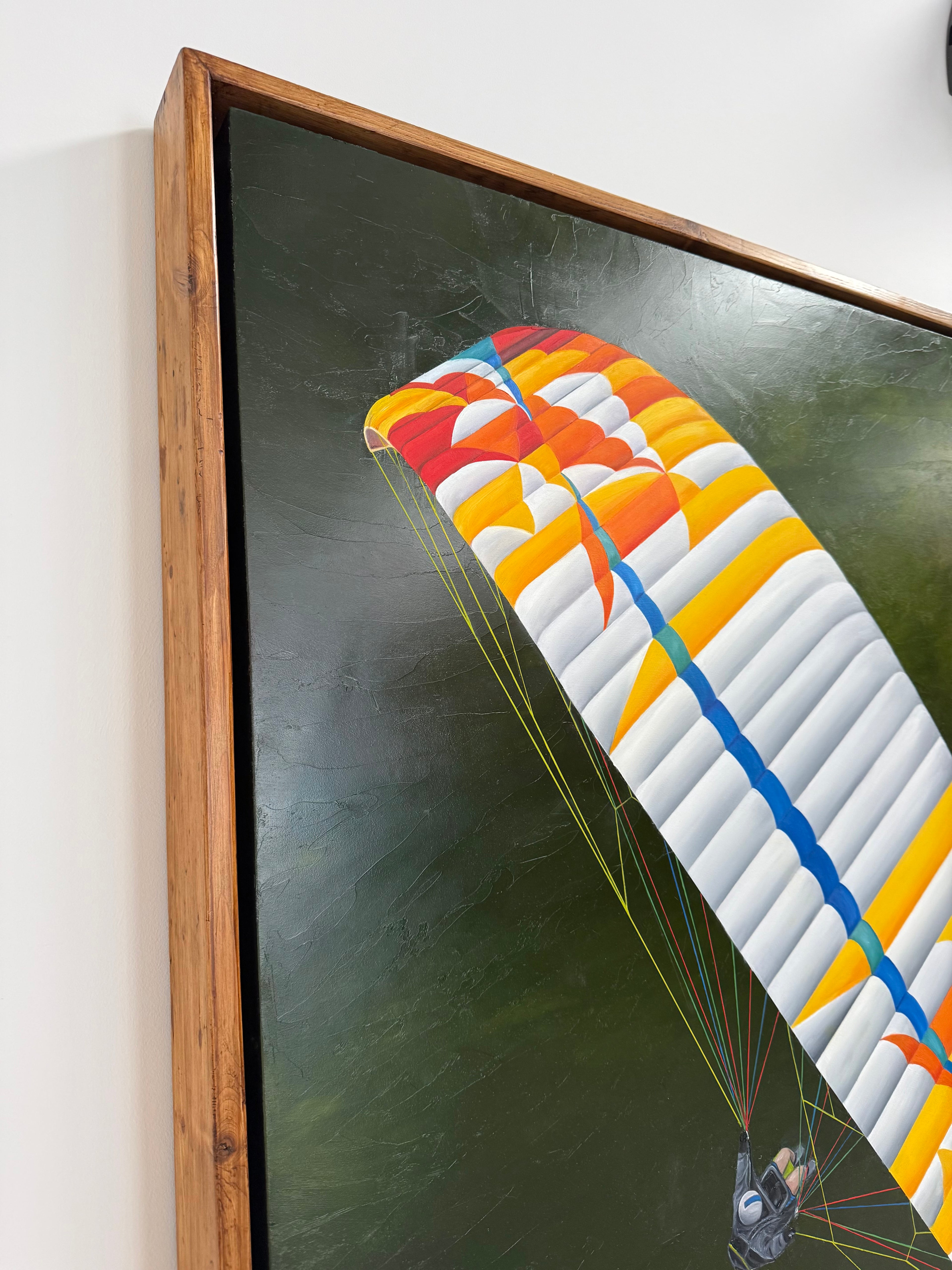 A framed artwork by katie hollinshead depicting a colorful paraglider canopy against a dark green background, shown hanging on a white wall. Handmade reclaimed wooden frame